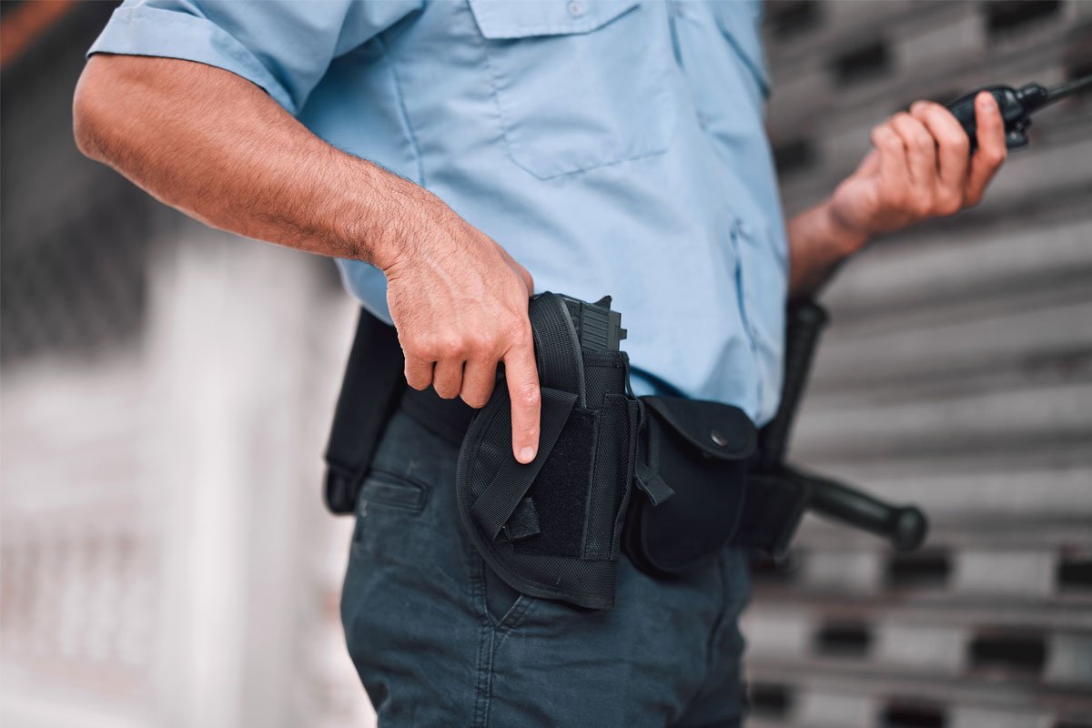 A security guard’s hand resting on a firearm in its holster, and they are also holding a handheld radio.