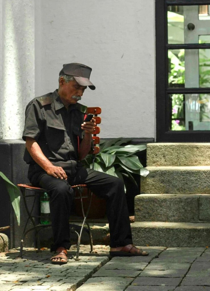 Man in uniform and cap sitting on a chair outdoors, looking at his phone.