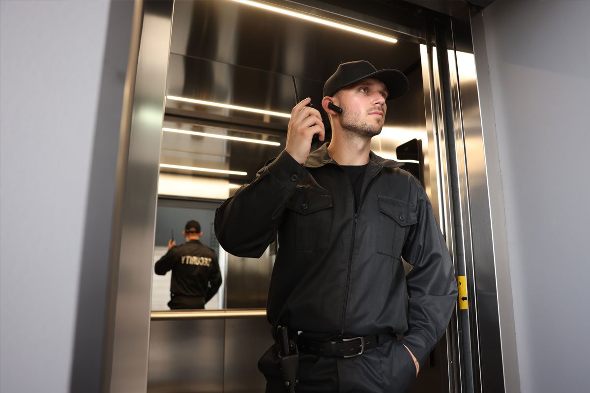 A male security guard standing inside a modern, metallic elevator.